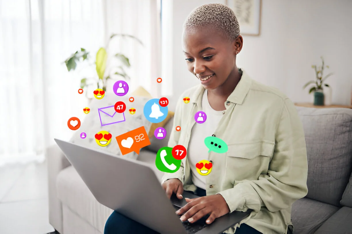 A smiling woman using a laptop at home with colorful social media notification icons floating around her, including likes, messages, and calls. The setting is bright and casual with plants in the background.